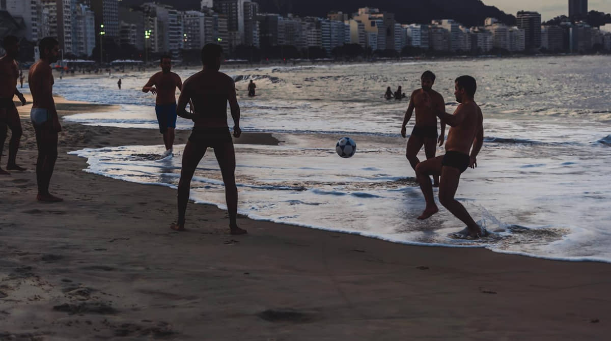 Homens jogam bola em praia no final da tarde